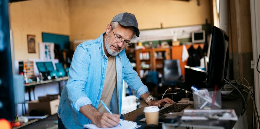 A person in a casual denim shirt and cap writes in a notebook while working at a desk with a computer and coffee in a workshop-style office.