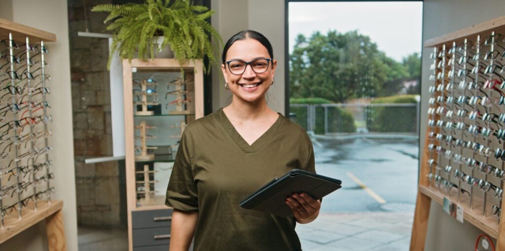 Person wearing a dark green scrub-style top holding a tablet in an optical store, surrounded by eyeglass display stands and indoor plants.