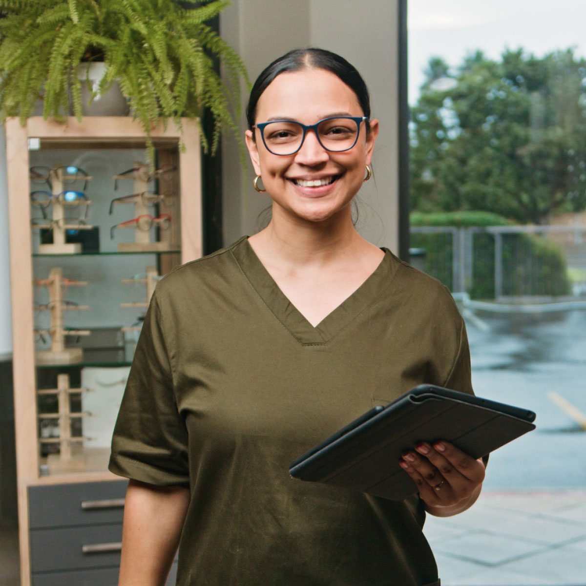 Person wearing a dark green scrub-style top holding a tablet in an optical store, surrounded by eyeglass display stands and indoor plants.