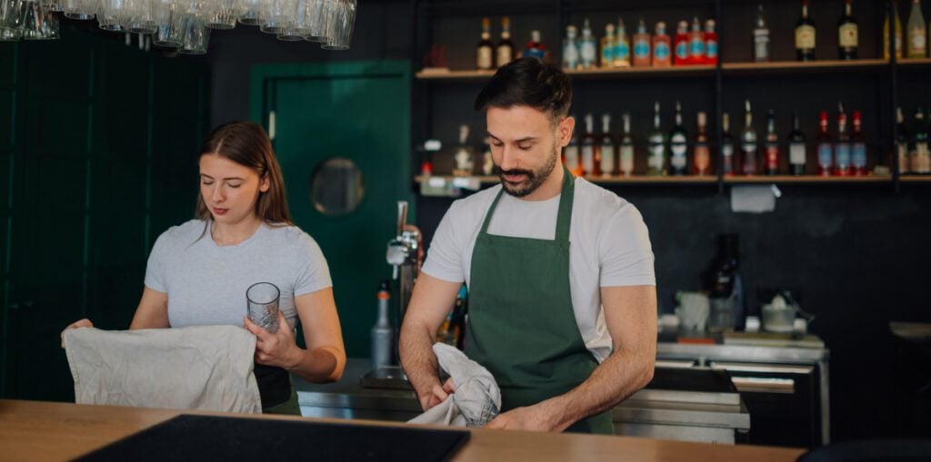 Two staff members preparing glasses and linens behind a bar.