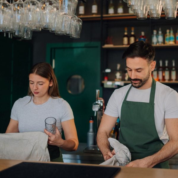 Two staff members preparing glasses and linens behind a bar.