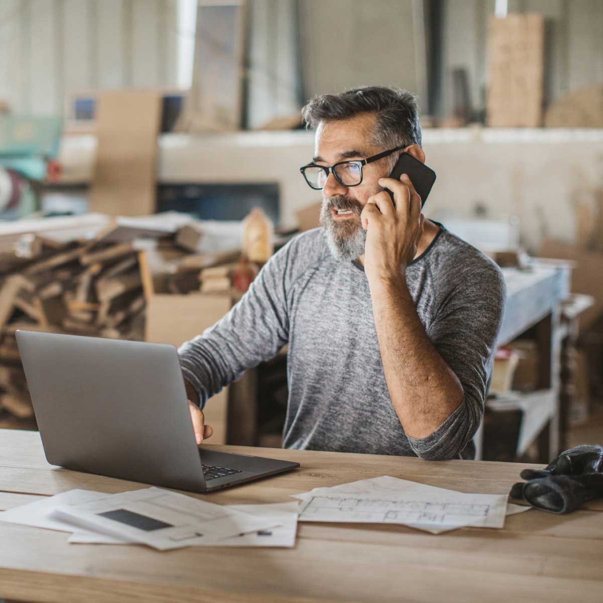 Carpenter on the phone while looking at a laptop in his workshop