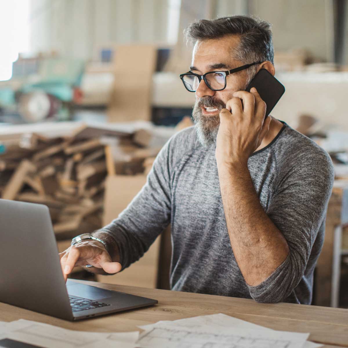 Person sitting at a workbench in a workshop, talking on a smartphone while using a laptop, with papers spread on the table and stacks of wooden materials in the background.