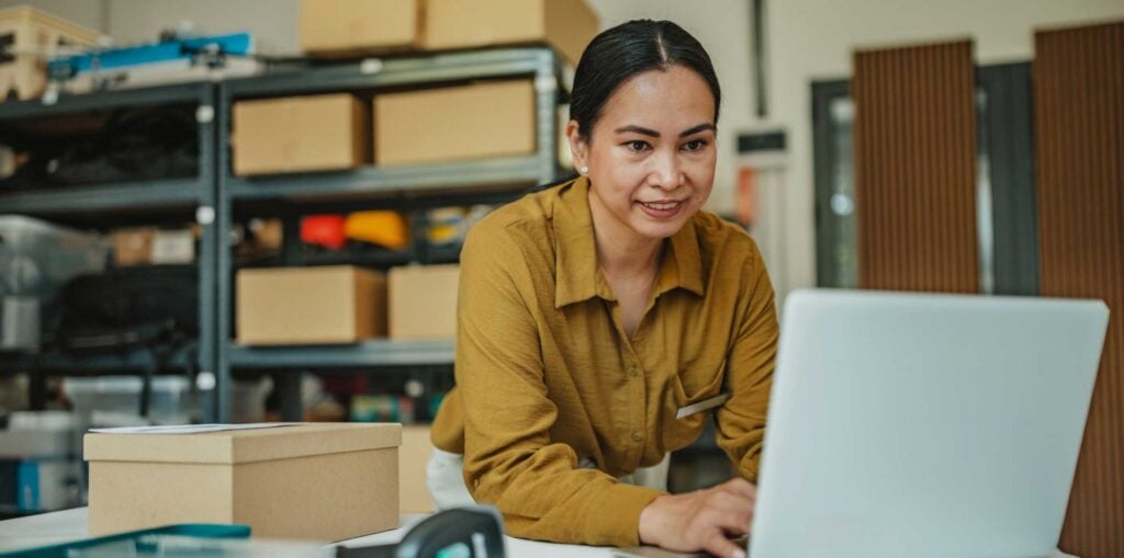 Small business owner working on a laptop at a packing table, surrounded by boxes and shelving in a warehouse workspace.