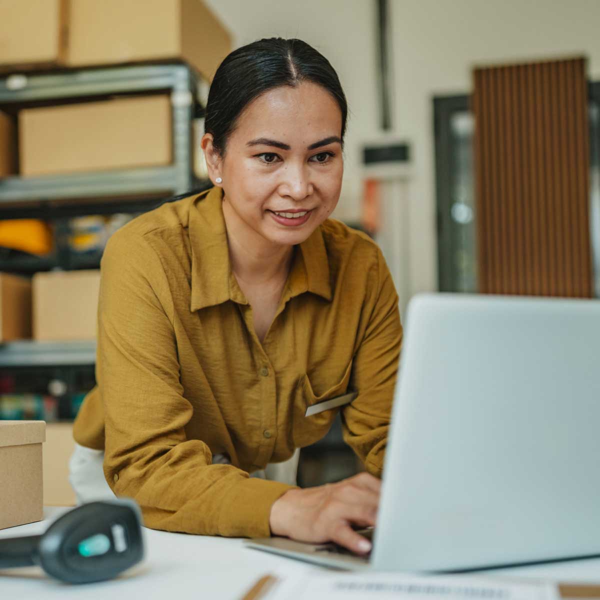 Small business owner working on a laptop at a packing table, surrounded by boxes and shelving in a warehouse workspace.