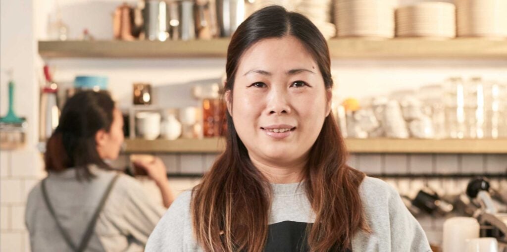 Cafe staff member standing at the counter holding a tablet, with another barista working in the background and shelves of cups and coffee equipment behind them.