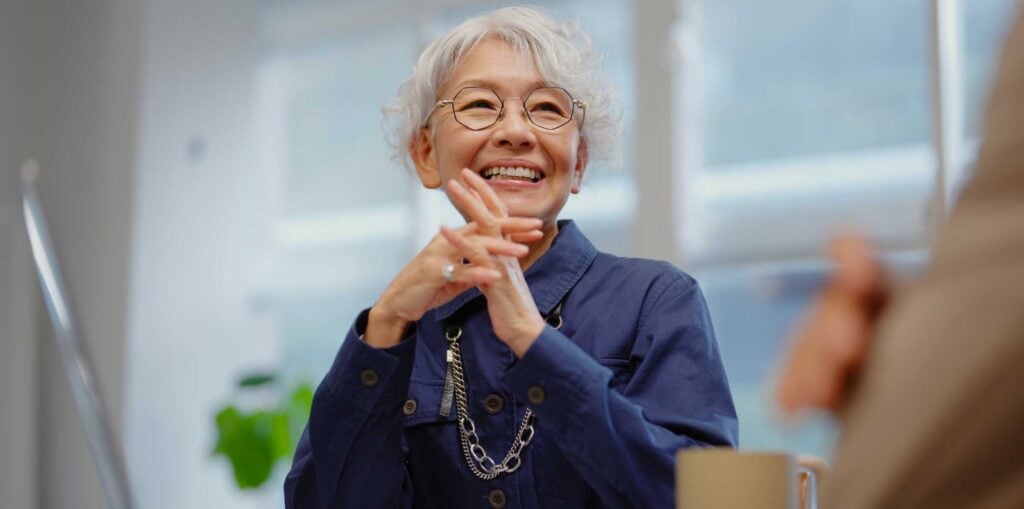 Person with short grey hair wearing a navy jacket and chain necklace, seated at a desk in a bright office, hands clasped while listening during a meeting
