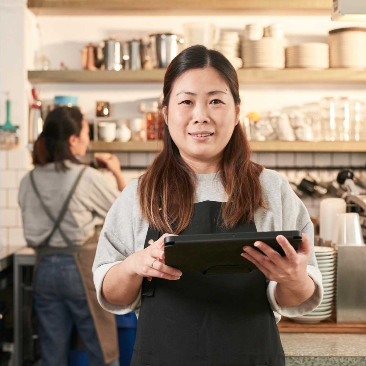 Cafe staff member standing at the counter holding a tablet, with another barista working in the background and shelves of cups and coffee equipment behind them.