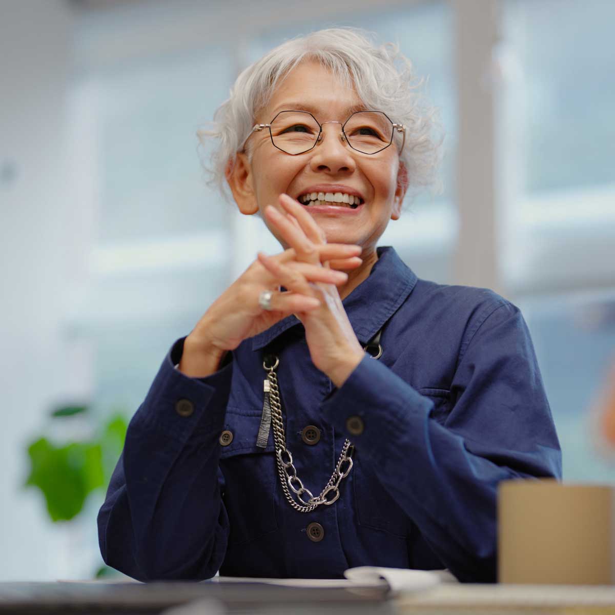 Person with short grey hair wearing a navy jacket and chain necklace, seated at a desk in a bright office, hands clasped while listening during a meeting