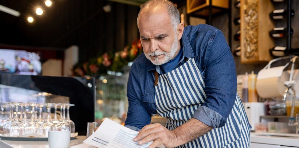 Small business owner in a café wearing a striped apron, reviewing paperwork at the counter with coffee equipment and shelves in the background.