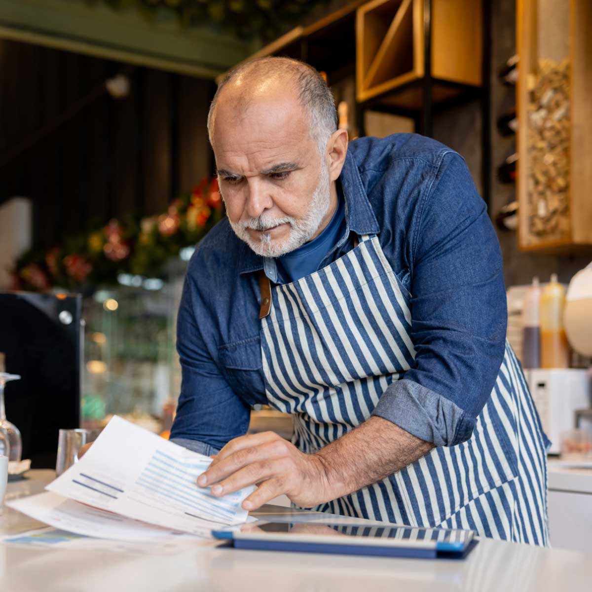 Small business owner in a café wearing a striped apron, reviewing paperwork at the counter with coffee equipment and shelves in the background.
