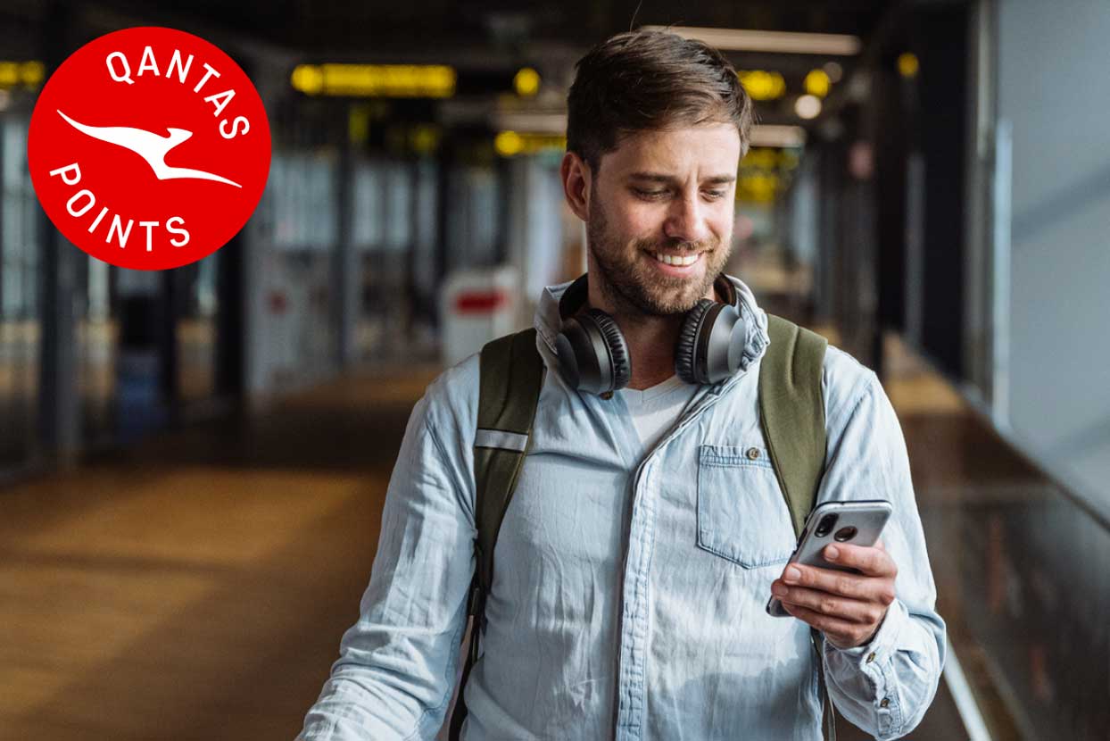 Traveller walking through an airport terminal with a backpack and headphones, checking a phone, alongside the Qantas Points logo.