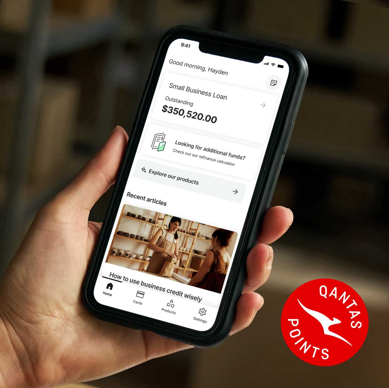 Close-up of a hand holding a smartphone displaying a small business loan app dashboard, showing an outstanding balance and recent articles. The phone is held in a warehouse setting, with a Qantas Points logo visible on the right