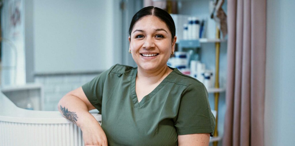 Person wearing green scrubs leaning against a counter in a clean, modern clinical setting with shelves of products in the background.