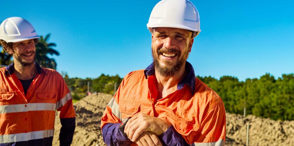 Construction worker wearing high‑visibility clothing and a hard hat, standing on a work site holding a tool.