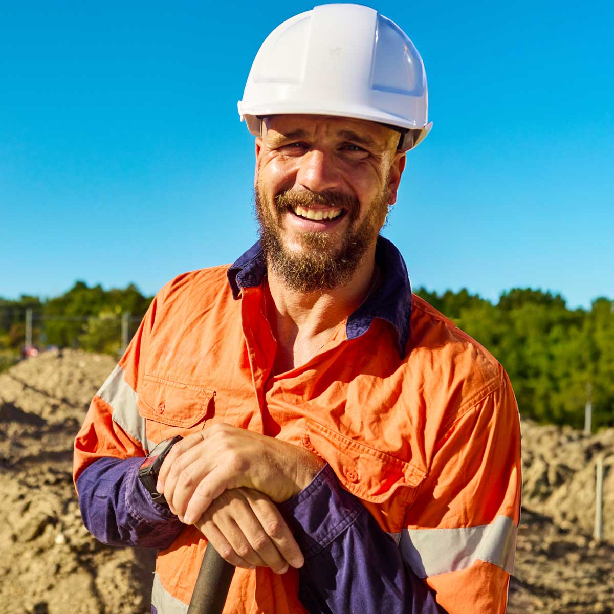 Construction worker wearing high‑visibility clothing and a hard hat, standing on a work site holding a tool.