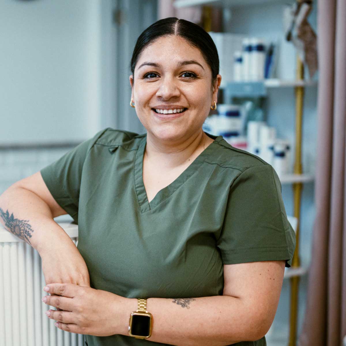 Person wearing green scrubs leaning against a counter in a clean, modern clinical setting with shelves of products in the background.