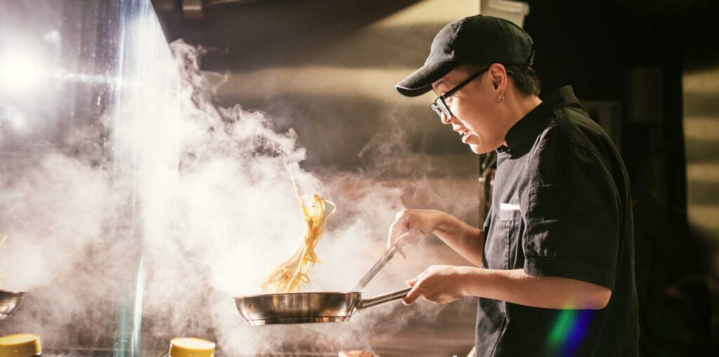 Chef in a commercial kitchen cooking over a stovetop, tossing noodles in a frying pan as steam rises.