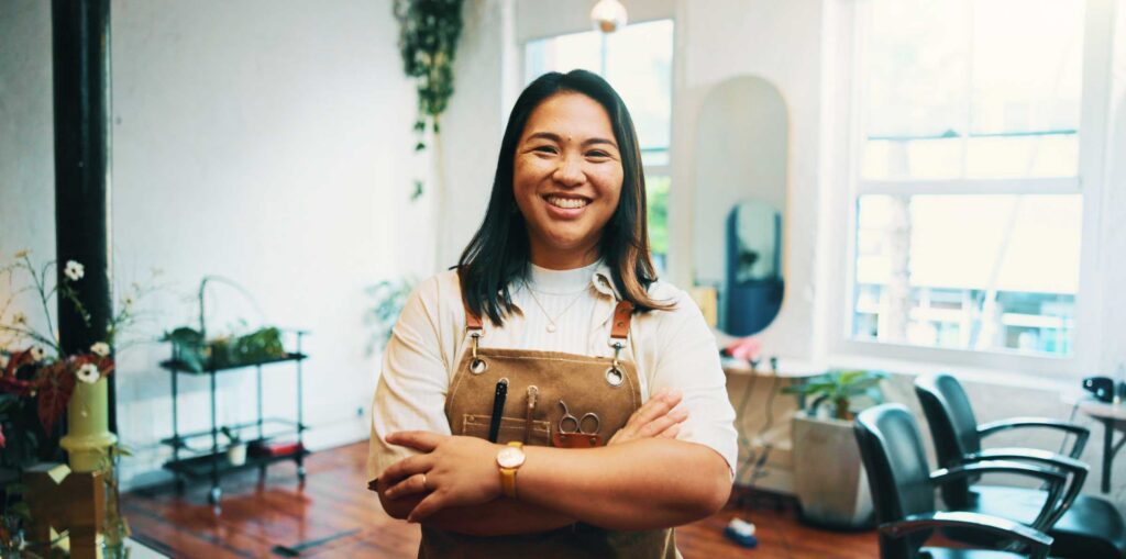 Small business owner standing with arms crossed in a hair salon, wearing an apron with tools, with styling chairs and plants in the background.
