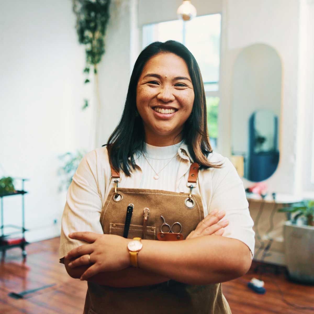 Small business owner standing with arms crossed in a hair salon, wearing an apron with tools, with styling chairs and plants in the background.