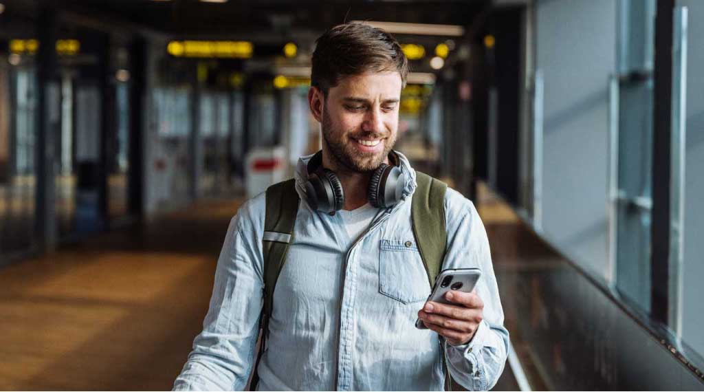 Traveller standing in an airport walkway, looking at a mobile phone while wearing headphones and a backpack.