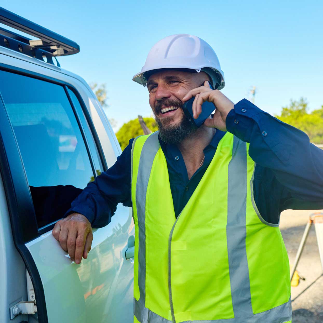 Tradesperson wearing a hi-vis vest and hard hat, standing by a work vehicle while talking on a mobile phone outdoors.
