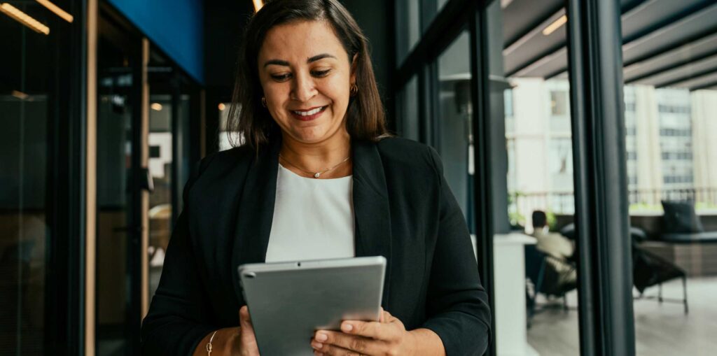Person in a modern office hallway using a tablet, with glass walls and workspaces in the background.