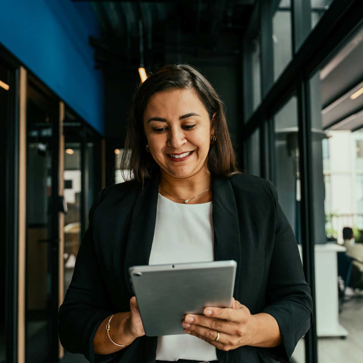 Person in a modern office hallway using a tablet, with glass walls and workspaces in the background.
