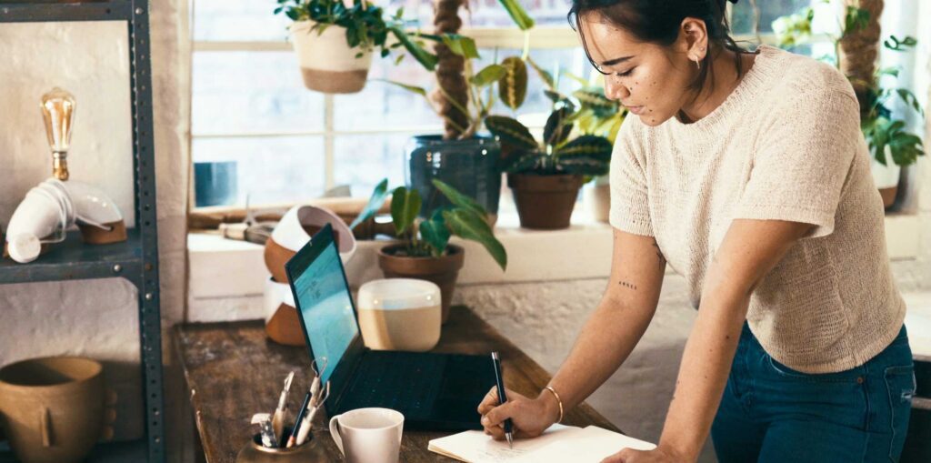 Person standing at a desk writing notes in a notebook beside a laptop, coffee cup and indoor plants.