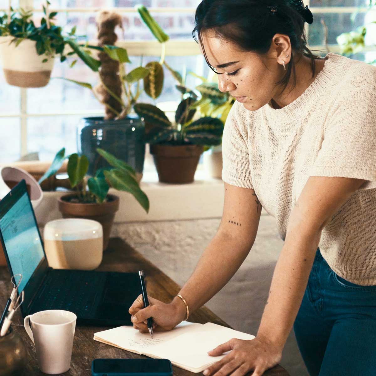 Person standing at a desk writing notes in a notebook beside a laptop, coffee cup and indoor plants.