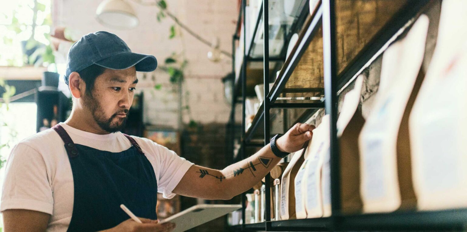Person in an apron checking inventory on a shelf while writing notes on a tablet in a small retail or café setting.