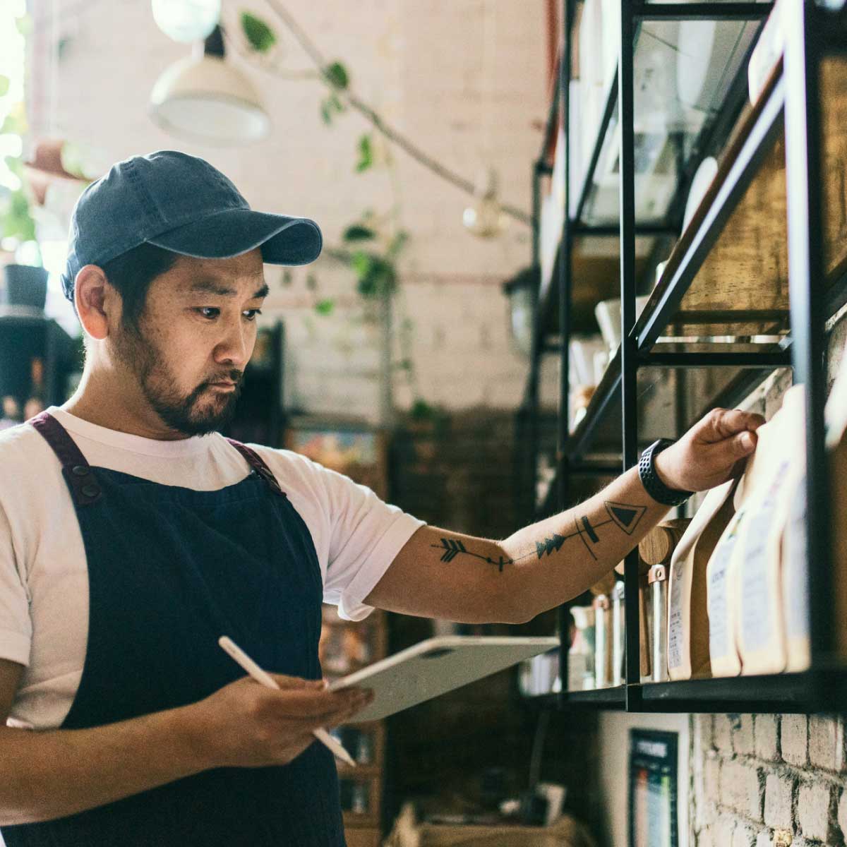 Person in an apron checking inventory on a shelf while writing notes on a tablet in a small retail or café setting.