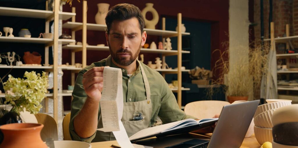 Small business owner reviewing receipts at a desk with a laptop, calculator and notebook in a workshop setting surrounded by ceramics and shelving.