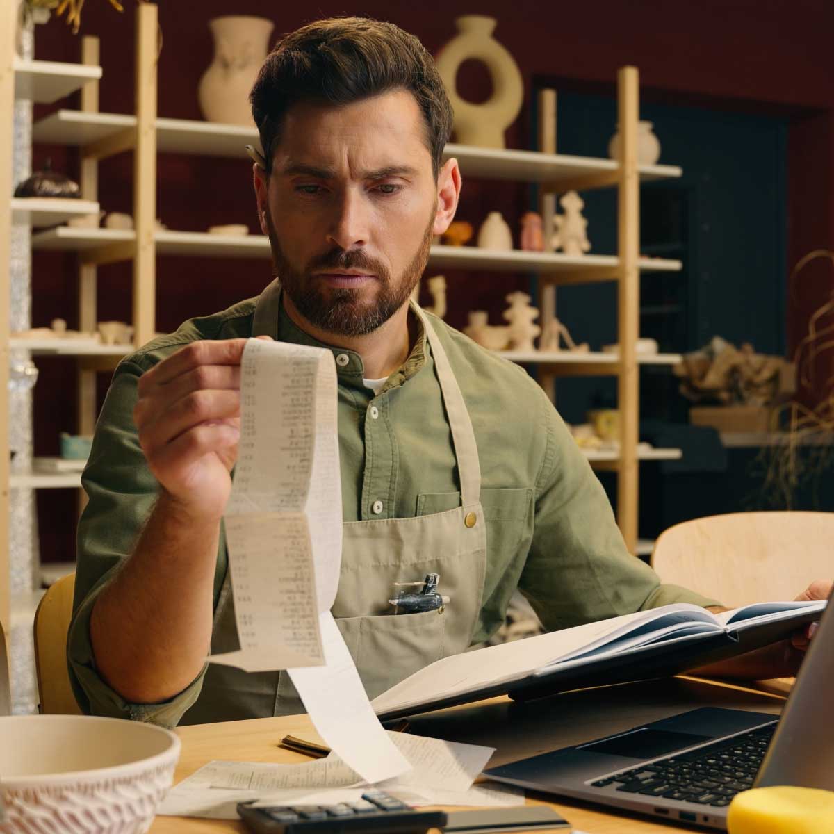 Small business owner reviewing receipts at a desk with a laptop, calculator and notebook in a workshop setting surrounded by ceramics and shelving.