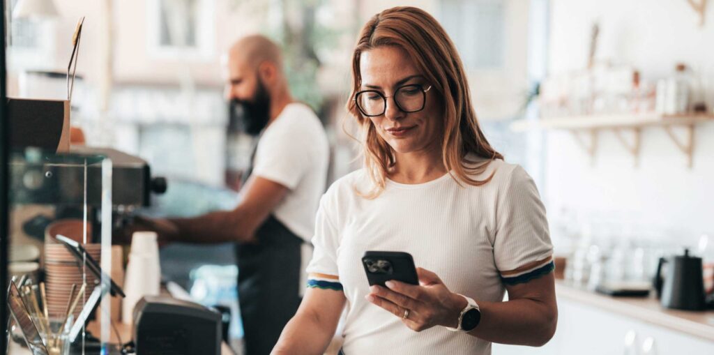 Person standing at a counter in a small business setting, looking at a smartphone while holding papers, with another person working on a laptop in the background.