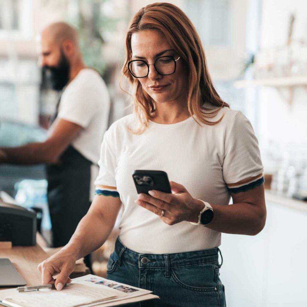 Person standing at a counter in a small business setting, looking at a smartphone while holding papers, with another person working on a laptop in the background.