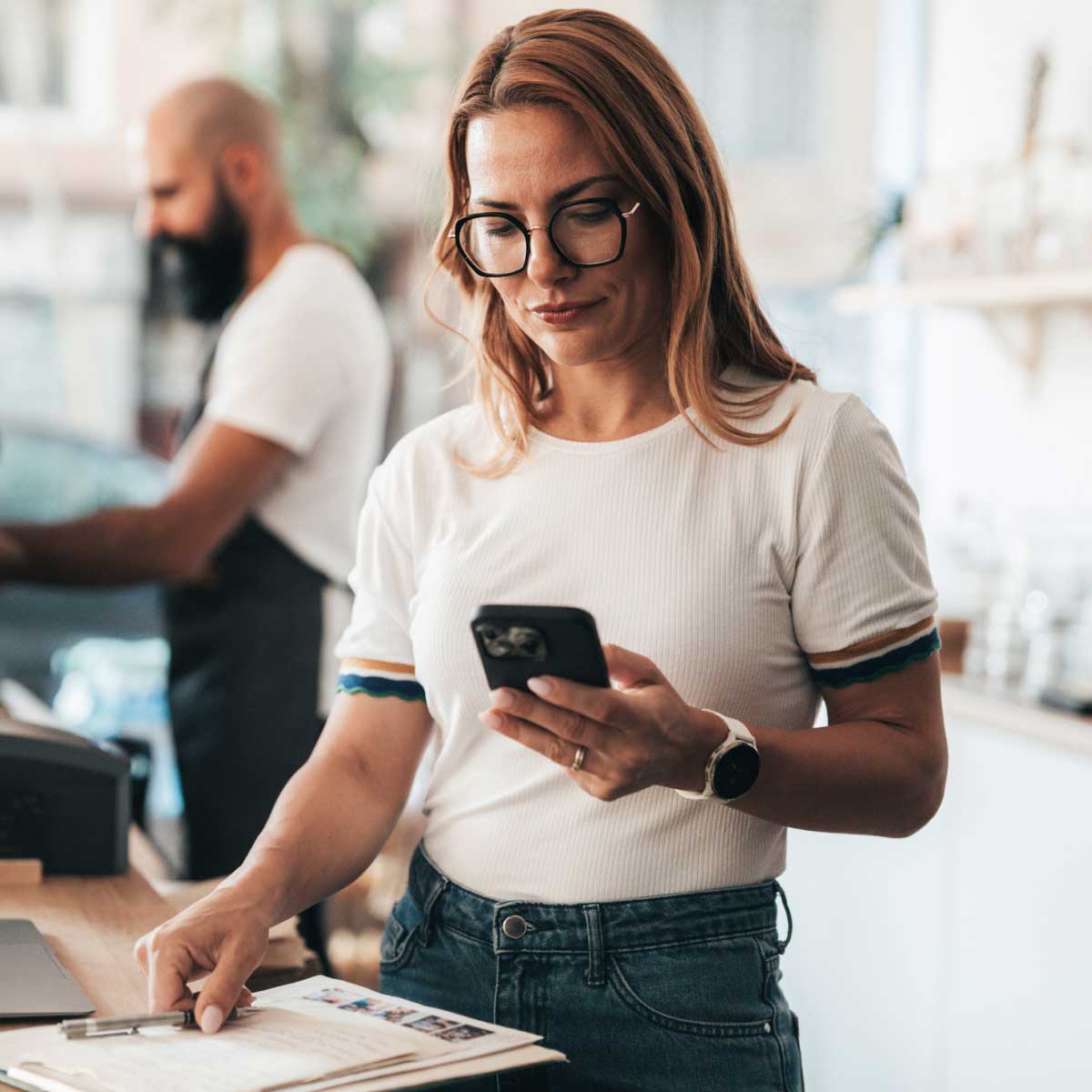 Person standing at a counter in a small business setting, looking at a smartphone while holding papers, with another person working on a laptop in the background.