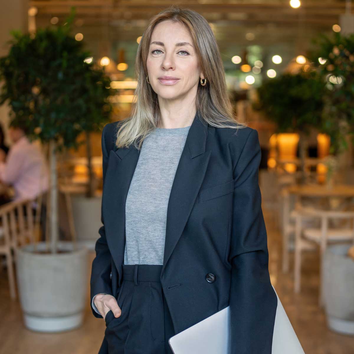 Businesslike confident woman in suit with laptop in hand standing in hall restaurant looks at camera stock photo