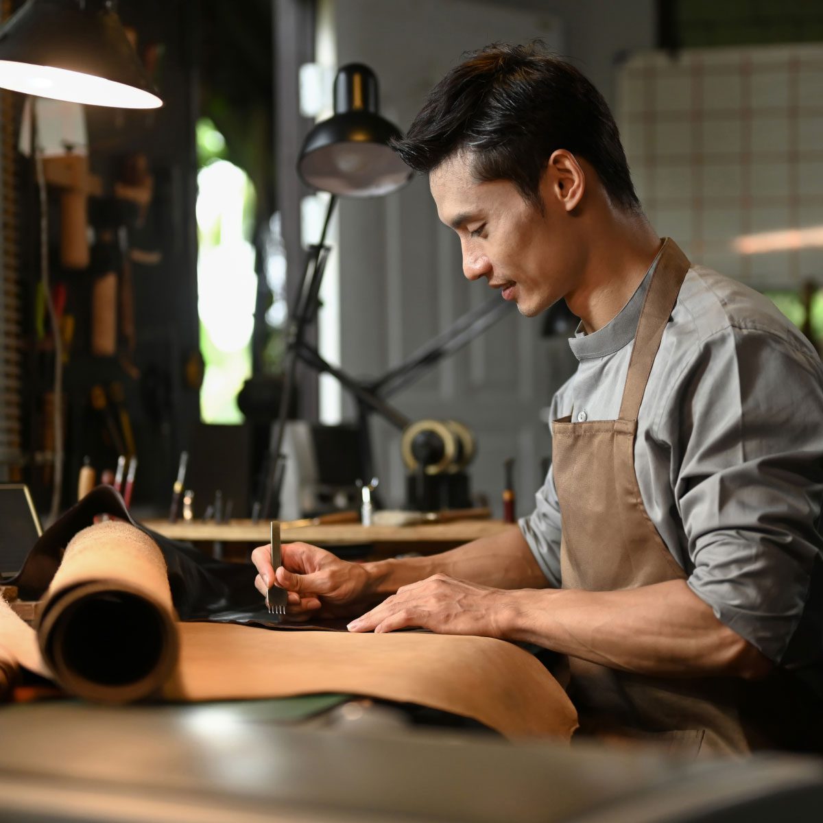 Person working in a leather workshop, using a stitching tool on a piece of leather at a workbench surrounded by rolled leather, tools and task lighting.