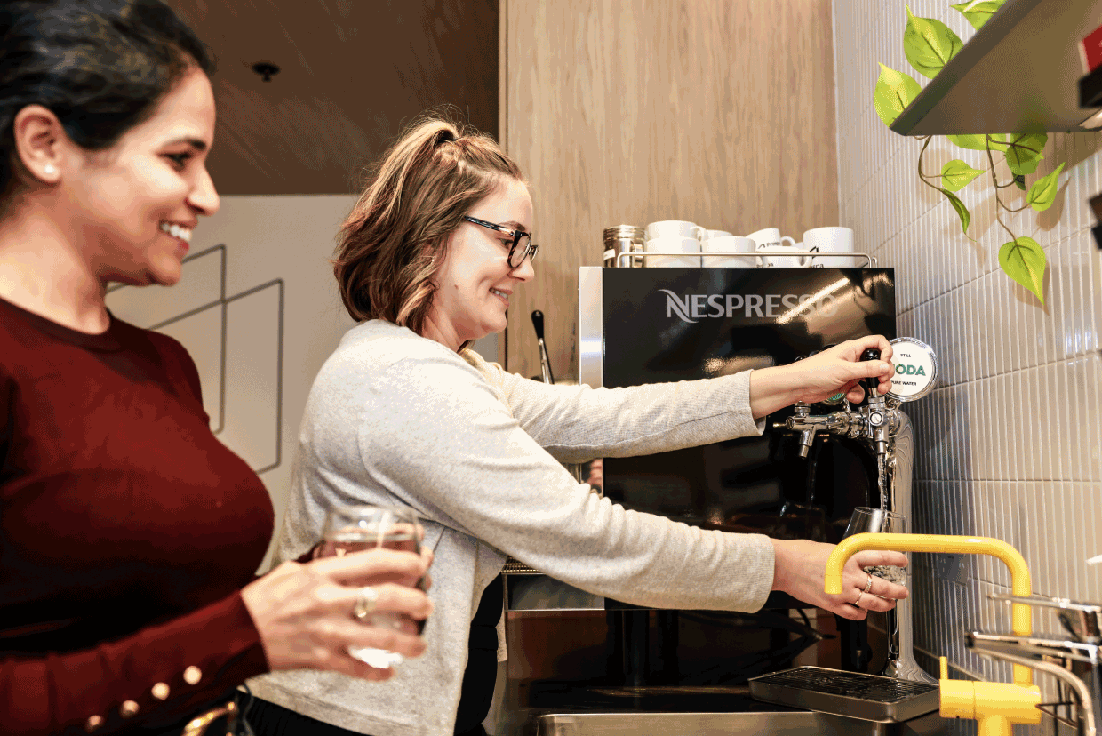 Two employees using a beverage station in an office kitchen area.
