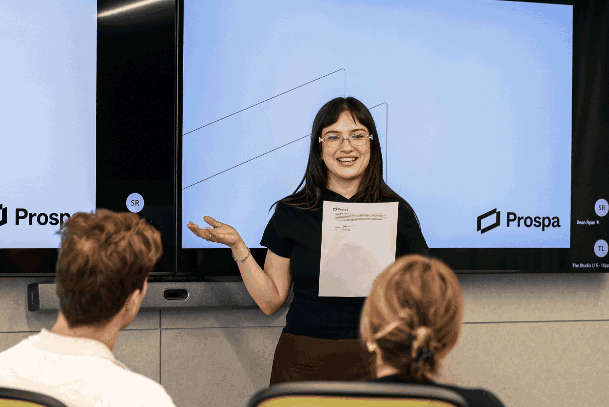 Employee presenting to colleagues in a meeting room with presentation screens displaying Prospa branding.