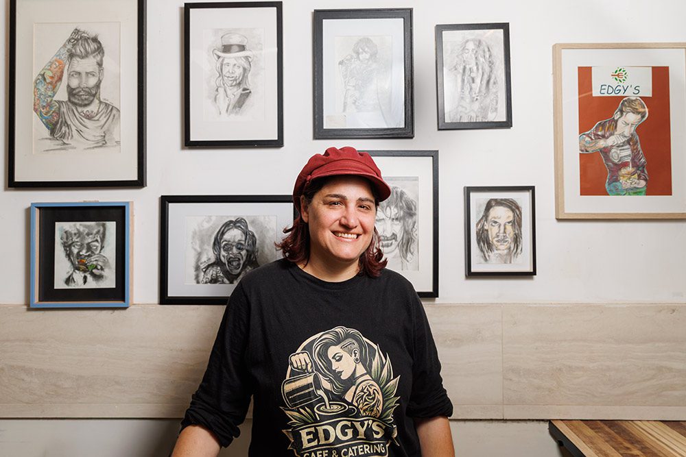 Maya Levy standing in Edgy's Cafe in front of a wall of framed portrait drawings she made