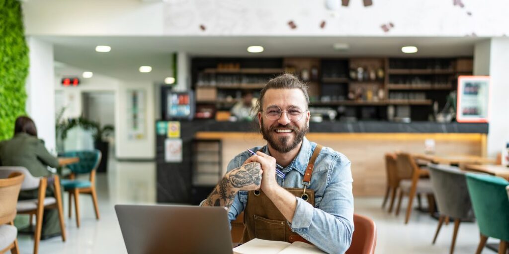 A smiling café owner in a denim shirt and leather apron sits at a wooden table with a laptop, notebook and phone. Hero image for the article on growing a business in stages.