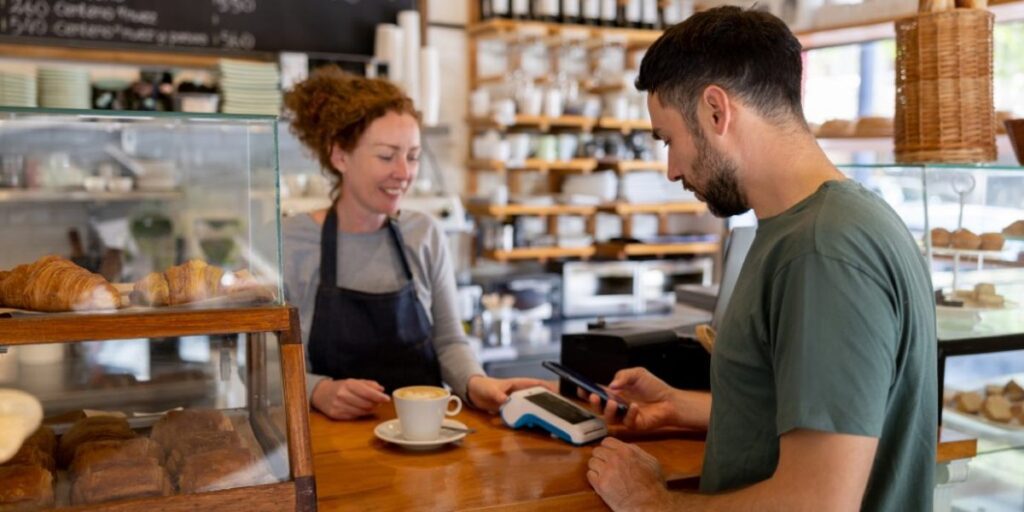 A smiling café worker serves a coffee while a customer pays by tapping his phone on the card terminal. Hero image for the article on the card surcharge ban.