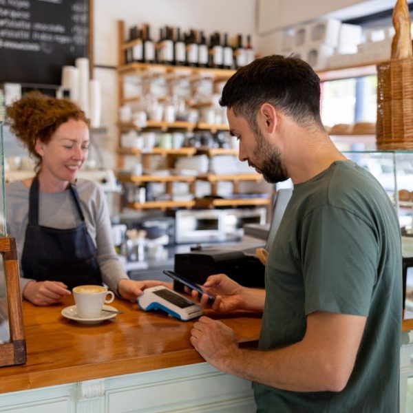 A smiling café worker serves a coffee while a customer pays by tapping his phone on the card terminal. Hero image for the article on the card surcharge ban.